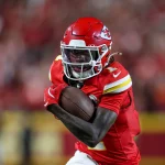 FootballR - NFL - Xavier Worthy - Chiefs besiegen Ravens - Ein Footballspieler in roter Uniform und Helm, der einen Football dicht am Körper hält, rennt entschlossen. Die Uniform des Spielers weist gelbe und weiße Akzente auf, im Hintergrund ist eine verschwommene Menschenmenge zu sehen. Das Logo auf seinem Helm zeigt, dass er Mitglied der Kansas City Chiefs ist, einem der Topteams der NFL. Diese Beschreibung wurde automatisch generiert.