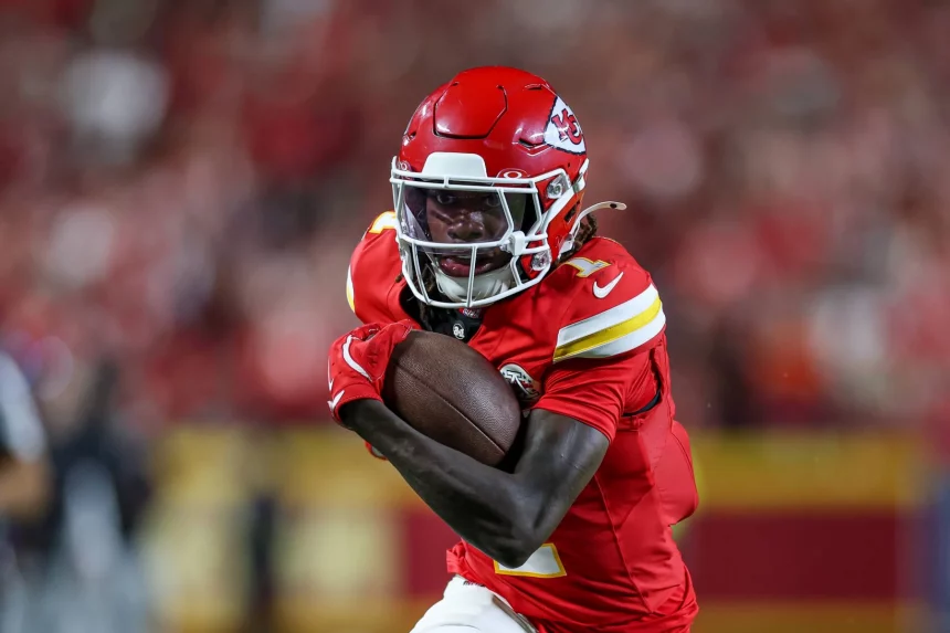 FootballR - NFL - Xavier Worthy - Chiefs besiegen Ravens - Ein Footballspieler in roter Uniform und Helm, der einen Football dicht am Körper hält, rennt entschlossen. Die Uniform des Spielers weist gelbe und weiße Akzente auf, im Hintergrund ist eine verschwommene Menschenmenge zu sehen. Das Logo auf seinem Helm zeigt, dass er Mitglied der Kansas City Chiefs ist, einem der Topteams der NFL. Diese Beschreibung wurde automatisch generiert.