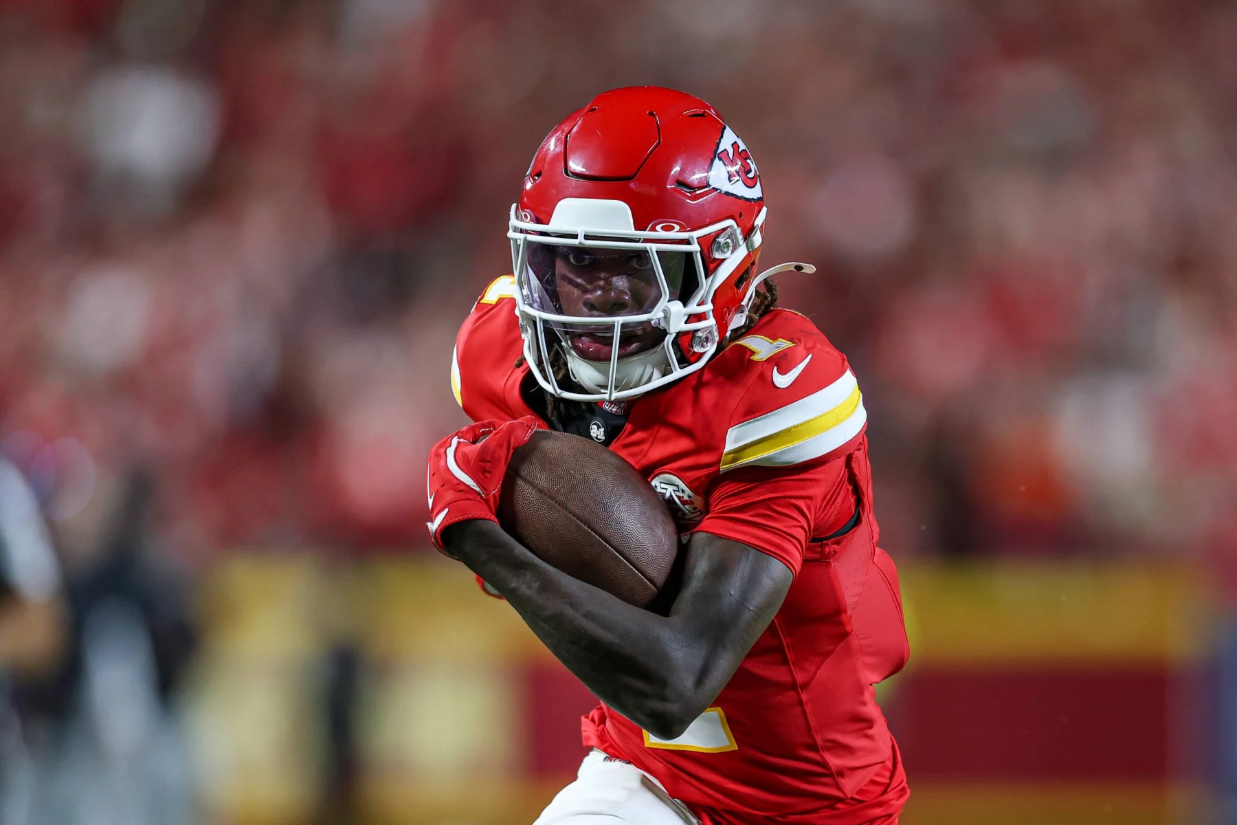 FootballR - NFL - Xavier Worthy - Chiefs besiegen Ravens - Ein Footballspieler in roter Uniform und Helm, der einen Football dicht am Körper hält, rennt entschlossen. Die Uniform des Spielers weist gelbe und weiße Akzente auf, im Hintergrund ist eine verschwommene Menschenmenge zu sehen. Das Logo auf seinem Helm zeigt, dass er Mitglied der Kansas City Chiefs ist, einem der Topteams der NFL. Diese Beschreibung wurde automatisch generiert.