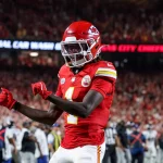 FootballR - NFL - Bye Week - Xavier Worthy - Ein Footballspieler in roter Uniform mit der Nummer 1 und „Chiefs“ auf seinem Trikot jubelt auf dem Feld. Er trägt einen roten Helm mit weißer Gesichtsmaske und schwarzen Handschuhen. Der unscharfe Hintergrund zeigt ein Stadion voller Fans und einige andere Spieler in weißen Uniformen, was unterstreicht, warum sie an der Spitze des NFL Power Ranking stehen. Diese Beschreibung wurde automatisch generiert.