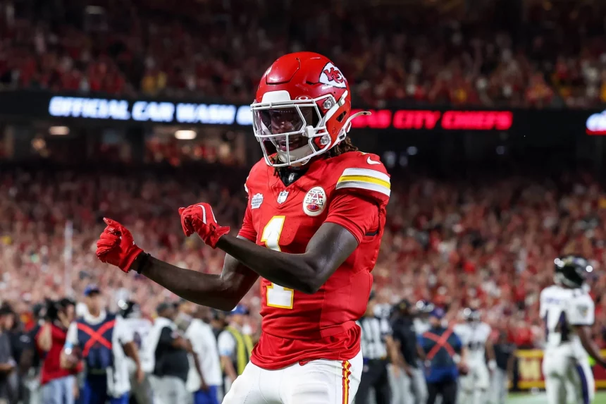 FootballR - NFL - Bye Week - Xavier Worthy - Ein Footballspieler in roter Uniform mit der Nummer 1 und „Chiefs“ auf seinem Trikot jubelt auf dem Feld. Er trägt einen roten Helm mit weißer Gesichtsmaske und schwarzen Handschuhen. Der unscharfe Hintergrund zeigt ein Stadion voller Fans und einige andere Spieler in weißen Uniformen, was unterstreicht, warum sie an der Spitze des NFL Power Ranking stehen. Diese Beschreibung wurde automatisch generiert.