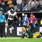 Eagles vs. Packers - NFL Saquon Barkley - Eagles-Spieler Nr. 26 springt während eines Spiels über einen am Boden liegenden Packers-Spieler und sorgt damit für eine ziemliche Spielfeld-Debatte. Die Action spielt sich auf dem Spielfeld ab, im Hintergrund sind Zuschauer und Veranstaltungspersonal zu sehen. Der Eagles-Spieler hält den Football fest, während der Packers-Spieler auf dem Rücken liegt und versucht, den Vorstoß zu stoppen. Spielfeld Debakel bei NFL Debüt in Brasilien. Diese Beschreibung wurde automatisch generiert.