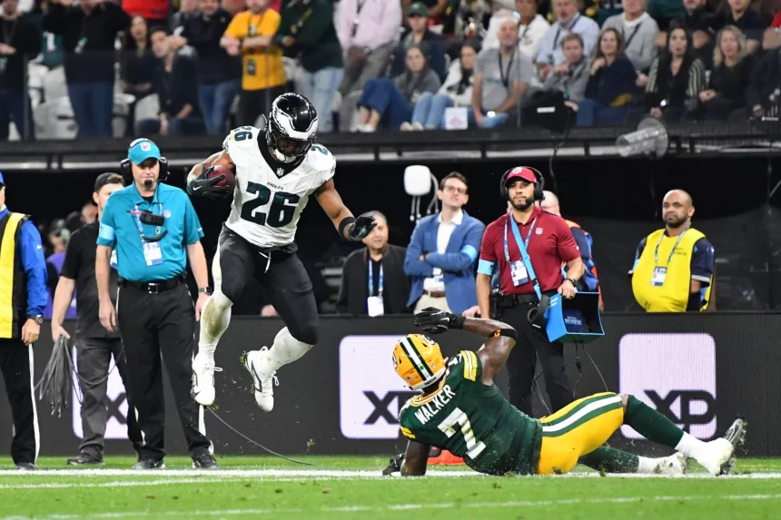 Eagles vs. Packers - NFL Saquon Barkley - Eagles-Spieler Nr. 26 springt während eines Spiels über einen am Boden liegenden Packers-Spieler und sorgt damit für eine ziemliche Spielfeld-Debatte. Die Action spielt sich auf dem Spielfeld ab, im Hintergrund sind Zuschauer und Veranstaltungspersonal zu sehen. Der Eagles-Spieler hält den Football fest, während der Packers-Spieler auf dem Rücken liegt und versucht, den Vorstoß zu stoppen. Spielfeld Debakel bei NFL Debüt in Brasilien. Diese Beschreibung wurde automatisch generiert.