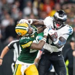 FootballR - NFL - Ein Footballspieler in weiß-grüner Uniform streckt beim Monday Night Football einen Spieler in grün-gelber Uniform mit ausgestreckten Armen aus. Der Spieler in Grün versucht, den gegnerischen Spieler A.J. Brown zu tackeln, wird dabei aber weggestoßen. Im Hintergrund ist eine Menge von Zuschauern und Offiziellen zu sehen. Diese Beschreibung wurde automatisch generiert.