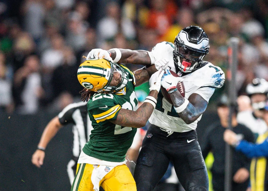 FootballR - NFL - Ein Footballspieler in weiß-grüner Uniform streckt beim Monday Night Football einen Spieler in grün-gelber Uniform mit ausgestreckten Armen aus. Der Spieler in Grün versucht, den gegnerischen Spieler A.J. Brown zu tackeln, wird dabei aber weggestoßen. Im Hintergrund ist eine Menge von Zuschauern und Offiziellen zu sehen. Diese Beschreibung wurde automatisch generiert.