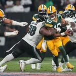 NFL Week 16 - NFL - Mehrere Footballspieler sind in ein Spiel verwickelt. In der Mitte wird Jordan Love, der eine grün-gelbe Uniform trägt und den Ball hält, von Gegnern in weiß-grünen Uniformen angegriffen. Andere Spieler beider Teams sind um sie herum in Aktion. Die Szene spielt auf einem Footballfeld mit einer sichtbaren Menschenmenge im Hintergrund. Diese Beschreibung wurde automatisch generiert.