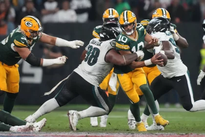 NFL Week 16 - NFL - Mehrere Footballspieler sind in ein Spiel verwickelt. In der Mitte wird Jordan Love, der eine grün-gelbe Uniform trägt und den Ball hält, von Gegnern in weiß-grünen Uniformen angegriffen. Andere Spieler beider Teams sind um sie herum in Aktion. Die Szene spielt auf einem Footballfeld mit einer sichtbaren Menschenmenge im Hintergrund. Diese Beschreibung wurde automatisch generiert.