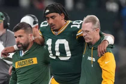 FootballR - NFL - Jordan Love - Ein Footballspieler in grün-gelber Uniform mit der Nummer 10 wird, flankiert von zwei Mitarbeitern, vom Feld geführt. Der Spieler hat zur Unterstützung einen Arm um jeden Mitarbeiter gelegt. Ein Mitarbeiter ist in Grün gekleidet, der andere in Gelb und Grün. Es scheint eine Schockmoment-Atmosphäre aus São Paulo zu sein. Diese Beschreibung wurde automatisch generiert.