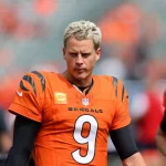 FootballR - NFL - Auf dem Spielfeld steht ein Footballspieler mit kurzen, hellblonden Haaren und einem orangefarbenen Trikot der Cincinnati Bengals mit der Nummer 9. Joe Burrows unverkennbares Selbstbewusstsein strahlt aus, er ist umgeben von seinen Teamkollegen in leuchtend orangefarbenen Uniformen. Im Hintergrund ist ein unscharfer Sitzbereich des Stadions zu sehen. Diese Beschreibung wurde automatisch generiert.