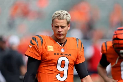 FootballR - NFL - Auf dem Spielfeld steht ein Footballspieler mit kurzen, hellblonden Haaren und einem orangefarbenen Trikot der Cincinnati Bengals mit der Nummer 9. Joe Burrows unverkennbares Selbstbewusstsein strahlt aus, er ist umgeben von seinen Teamkollegen in leuchtend orangefarbenen Uniformen. Im Hintergrund ist ein unscharfer Sitzbereich des Stadions zu sehen. Diese Beschreibung wurde automatisch generiert.