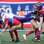 FootballR - NFL - Ein Footballspieler in einer weiß-lila Uniform wird in die Luft geschleudert, nachdem er während eines Spiels der NFL Saison 2024 von einem Spieler in einer rot-blauen Uniform angegriffen wurde. Ein weiterer Spieler in Rot und Blau ist in der Nähe, im Hintergrund ist ein Schiedsrichter inmitten des Geschehens auf dem Spielfeld zu sehen. Die Zuschauermenge ist nicht zu sehen. Diese Beschreibung wurde automatisch generiert.