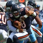 FootballR - NFL - Caleb Williams - Ein Footballspieler in schwarzer Uniform mit der Nummer 18, der den Ball hält, wird von einem Gegner in weiß-blauer Uniform angegriffen. Ein weiterer Spieler in ähnlicher schwarzer Uniform mit der Nummer 20 ist links zu sehen. Im Hintergrund auf dem Spielfeld sind weitere Spieler in blauen Uniformen zu sehen, was an eine klassische Szene der Chicago Bears erinnert. Diese Beschreibung wurde automatisch generiert.