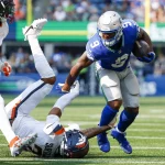 FootballR - NFL - Kenneth Walker, ein Footballspieler in blau-silberner Uniform, rennt mit dem Ball und wehrt einen Verteidiger der gegnerischen Mannschaft ab, der in weiß-orangefarbener Uniform gekleidet ist. Der Verteidiger fällt rückwärts auf das Spielfeld. Ein weiterer Spieler in weiß-orangefarbener Uniform ist im linken Hintergrund teilweise sichtbar. Diese Beschreibung wurde automatisch generiert.