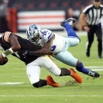 FootballR - NFL - Ein Footballspieler der Cleveland Browns, Deshaun Watson, in brauner Uniform wird von einem Spieler der Dallas Cowboys in weiß-blauer Uniform angegriffen und fällt zu Boden. Grüner Rasen ist zu sehen, im Hintergrund sind Schiedsrichter in schwarz-weißen Streifen zu sehen. Diese Beschreibung wurde automatisch generiert.