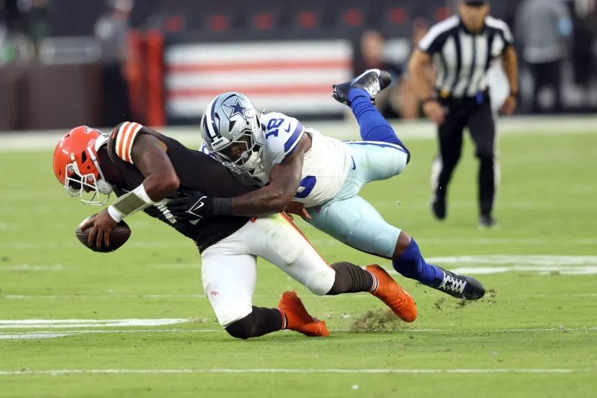 FootballR - NFL - Ein Footballspieler der Cleveland Browns, Deshaun Watson, in brauner Uniform wird von einem Spieler der Dallas Cowboys in weiß-blauer Uniform angegriffen und fällt zu Boden. Grüner Rasen ist zu sehen, im Hintergrund sind Schiedsrichter in schwarz-weißen Streifen zu sehen. Diese Beschreibung wurde automatisch generiert.