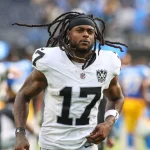 FootballR - NFL - Davante Adams, der eine weiß-schwarze Uniform mit der Nummer 17 und einem LV-Aufnäher trägt, läuft über das Feld. Mit langen Dreadlocks, einem schwarzen Stirnband und sichtbaren Tattoos auf den Armen fällt er vor den verschwommenen Figuren und den Stadionsitzen in Blau- und Gelbtönen auf. Diese Beschreibung wurde automatisch generiert.