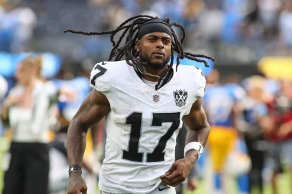FootballR - NFL - Davante Adams, der eine weiß-schwarze Uniform mit der Nummer 17 und einem LV-Aufnäher trägt, läuft über das Feld. Mit langen Dreadlocks, einem schwarzen Stirnband und sichtbaren Tattoos auf den Armen fällt er vor den verschwommenen Figuren und den Stadionsitzen in Blau- und Gelbtönen auf. Diese Beschreibung wurde automatisch generiert.