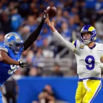 FootballR - NFL - In einem Moment eines Footballspiels wirft ein Quarterback der Rams in weißem Trikot und gelber Hose den Ball. Marcus Davenport, ein Lions-Spieler in blauem Trikot und silbernem Helm, versucht, den Pass zu blockieren. Im Hintergrund ist eine verschwommene Zuschauermenge im Stadion zu sehen. Diese Beschreibung wurde automatisch generiert.