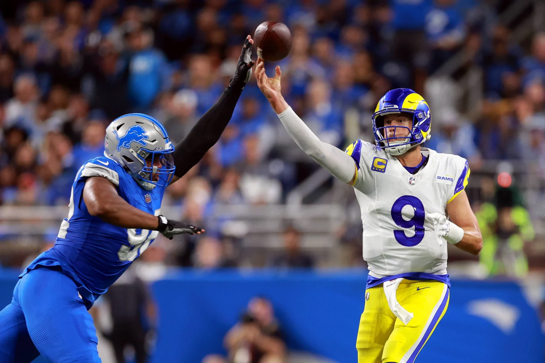 FootballR - NFL - In einem Moment eines Footballspiels wirft ein Quarterback der Rams in weißem Trikot und gelber Hose den Ball. Marcus Davenport, ein Lions-Spieler in blauem Trikot und silbernem Helm, versucht, den Pass zu blockieren. Im Hintergrund ist eine verschwommene Zuschauermenge im Stadion zu sehen. Diese Beschreibung wurde automatisch generiert.