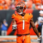 FootballR - NFL - Ein Footballspieler in einer orangefarbenen Cincinnati Bengals Uniform mit schwarzen Streifen und der Nummer 1 auf der Brust steht auf dem Feld und zeigt nach vorne. Der Helm des Spielers passt zur Uniform. Im Hintergrund sind Zuschauer und Offizielle unscharf zu sehen, was darauf hindeutet, dass ein Spiel im Gange ist, bei dem Ja'Marr Chase die Führung übernimmt. Diese Beschreibung wurde automatisch generiert.