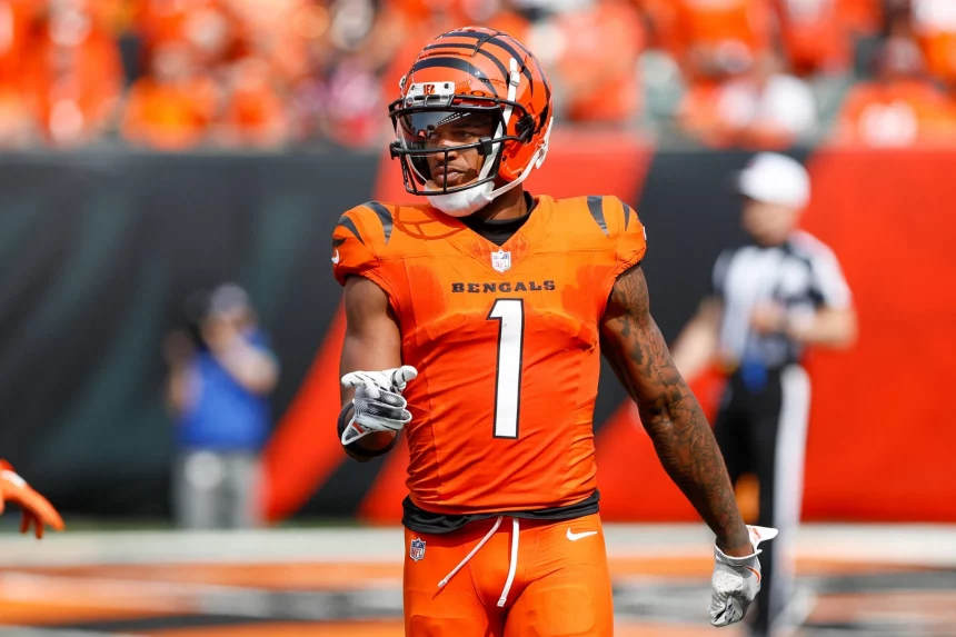 FootballR - NFL - Ein Footballspieler in einer orangefarbenen Cincinnati Bengals Uniform mit schwarzen Streifen und der Nummer 1 auf der Brust steht auf dem Feld und zeigt nach vorne. Der Helm des Spielers passt zur Uniform. Im Hintergrund sind Zuschauer und Offizielle unscharf zu sehen, was darauf hindeutet, dass ein Spiel im Gange ist, bei dem Ja'Marr Chase die Führung übernimmt. Diese Beschreibung wurde automatisch generiert.