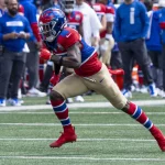 FootballR - NFL - Ein Footballspieler in blau-rot-weißer Uniform mit goldenen Hosen und roten Stollen läuft mitten auf dem Feld. Mit Helm und Visier nimmt er eine Actionpose ein, als würde er rennen oder sich auf einen Tackling vorbereiten. Im Hintergrund stehen Teamkollegen und Mitarbeiter in blauer Kleidung an der Seitenlinie. Es könnte Malik Nabers in Bewegung sein. Diese Beschreibung wurde automatisch generiert.