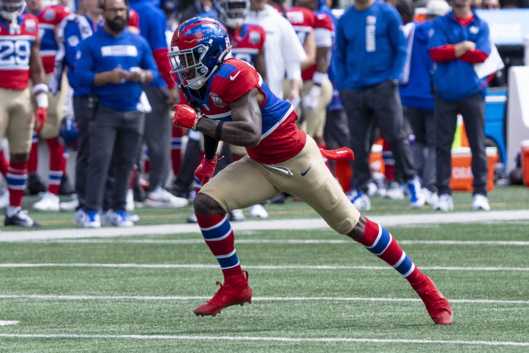 FootballR - NFL - Ein Footballspieler in blau-rot-weißer Uniform mit goldenen Hosen und roten Stollen läuft mitten auf dem Feld. Mit Helm und Visier nimmt er eine Actionpose ein, als würde er rennen oder sich auf einen Tackling vorbereiten. Im Hintergrund stehen Teamkollegen und Mitarbeiter in blauer Kleidung an der Seitenlinie. Es könnte Malik Nabers in Bewegung sein. Diese Beschreibung wurde automatisch generiert.