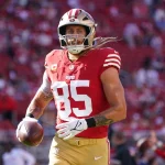 FootballR - NFL Fantasy Football Woche 8 - George Kittle - Ein Footballspieler in einem roten Trikot mit der Nummer 85 und einem goldenen Helm läuft mit einem Football in der Hand über das Feld. Das Trikot hat weiße Streifen an den Ärmeln und Mannschaftswappen auf Brust und Schultern. Im Hintergrund ist ein Stadion voller Zuschauer zu sehen, was an George Kittle erinnert, der für die 49ers spielte. Diese Beschreibung wurde automatisch generiert.