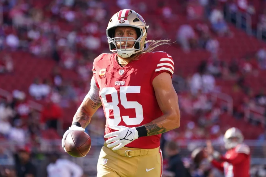 FootballR - NFL Fantasy Football Woche 8 - George Kittle - Ein Footballspieler in einem roten Trikot mit der Nummer 85 und einem goldenen Helm läuft mit einem Football in der Hand über das Feld. Das Trikot hat weiße Streifen an den Ärmeln und Mannschaftswappen auf Brust und Schultern. Im Hintergrund ist ein Stadion voller Zuschauer zu sehen, was an George Kittle erinnert, der für die 49ers spielte. Diese Beschreibung wurde automatisch generiert.