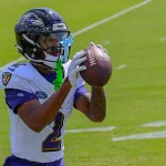 FootballR - NFL - Ein Ravens-Rookie-Footballspieler, Nate Wiggins, trägt ein weiß-lila Baltimore Ravens-Trikot und einen Helm und fängt einen Football. An seiner Gesichtsmaske sind grün-blaue Mundschützer befestigt. Der Hintergrund zeigt ein grünes Feld mit weißen Yard-Linien während des Spiels gegen Raiders. Diese Beschreibung wurde automatisch generiert.
