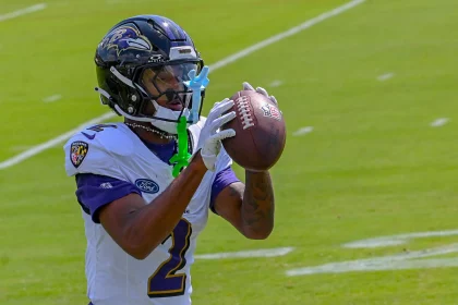 FootballR - NFL - Ein Ravens-Rookie-Footballspieler, Nate Wiggins, trägt ein weiß-lila Baltimore Ravens-Trikot und einen Helm und fängt einen Football. An seiner Gesichtsmaske sind grün-blaue Mundschützer befestigt. Der Hintergrund zeigt ein grünes Feld mit weißen Yard-Linien während des Spiels gegen Raiders. Diese Beschreibung wurde automatisch generiert.