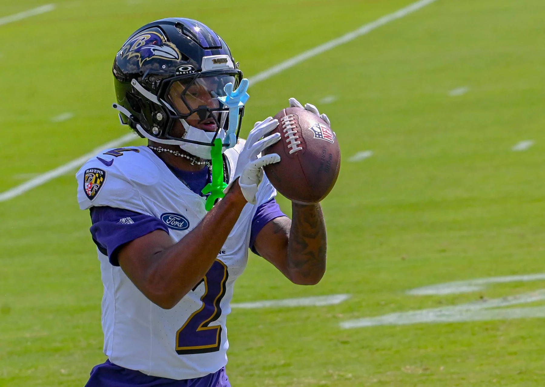 FootballR - NFL - Ein Ravens-Rookie-Footballspieler, Nate Wiggins, trägt ein weiß-lila Baltimore Ravens-Trikot und einen Helm und fängt einen Football. An seiner Gesichtsmaske sind grün-blaue Mundschützer befestigt. Der Hintergrund zeigt ein grünes Feld mit weißen Yard-Linien während des Spiels gegen Raiders. Diese Beschreibung wurde automatisch generiert.