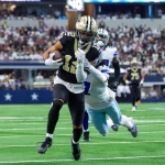 FootballR - NFL - Chris Olave - Ein Footballspieler in schwarz-goldener Uniform läuft während eines Falcons-Spiels mit dem Ball, verfolgt von einem gegnerischen Spieler in weiß-blauer Uniform. Sie spielen auf einem grünen Feld, hinter ihnen eine Menge Zuschauer auf den Tribünen. Die Szene ist actionreich und intensiv. Diese Beschreibung wurde automatisch generiert.