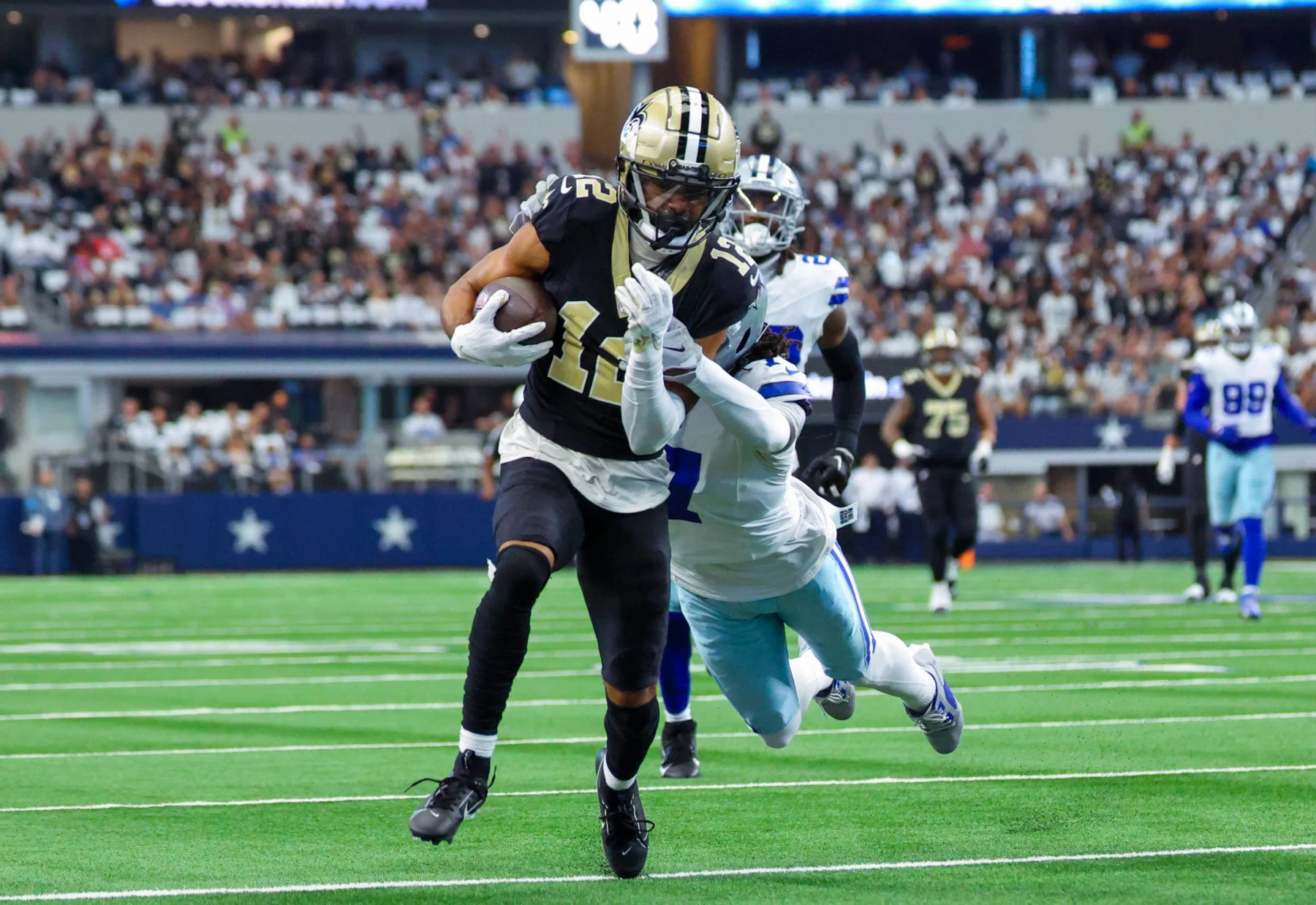 FootballR - NFL - Chris Olave - Ein Footballspieler in schwarz-goldener Uniform läuft während eines Falcons-Spiels mit dem Ball, verfolgt von einem gegnerischen Spieler in weiß-blauer Uniform. Sie spielen auf einem grünen Feld, hinter ihnen eine Menge Zuschauer auf den Tribünen. Die Szene ist actionreich und intensiv. Diese Beschreibung wurde automatisch generiert.