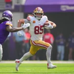 FootballR - NFL - George Kittle - Ein Footballspieler in weiß-roter Uniform mit der Nummer 85 rennt mit dem Ball nach vorne, während ihn ein Spieler in lila-weißer Uniform verfolgt. Die Szene spielt sich während eines Spiels auf einem Fußballfeld ab. Diese Beschreibung wurde automatisch generiert.