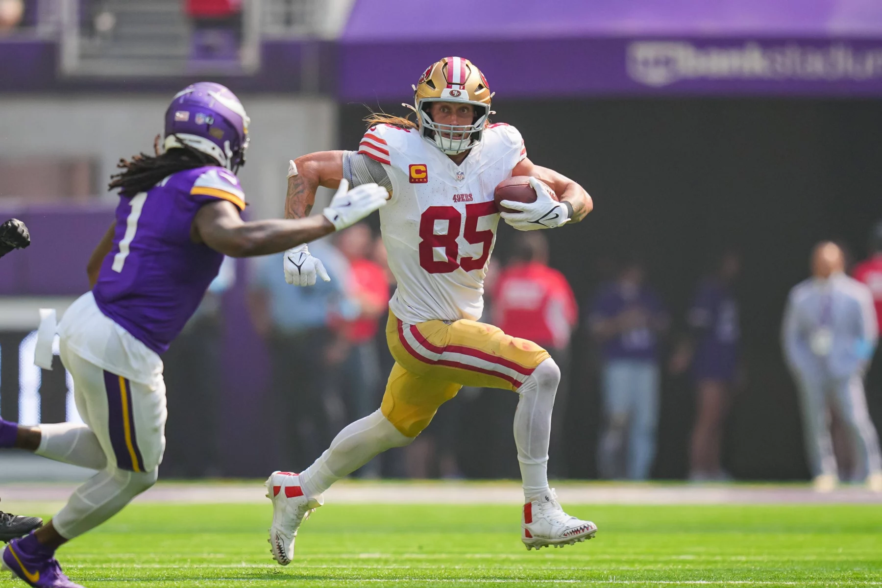 FootballR - NFL - George Kittle - Ein Footballspieler in weiß-roter Uniform mit der Nummer 85 rennt mit dem Ball nach vorne, während ihn ein Spieler in lila-weißer Uniform verfolgt. Die Szene spielt sich während eines Spiels auf einem Fußballfeld ab. Diese Beschreibung wurde automatisch generiert.