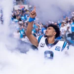 FootballR - NFL - Bryce Young - Ein Footballspieler trägt ein weißes Panthers-Trikot mit der Nummer 9 und blauen und schwarzen Akzenten und ist von Rauch umgeben, während er mit seiner rechten Hand nach oben gestikuliert. Er steht auf einem Spielfeld und die Tribünen im Hintergrund sind voller Fans, von denen einige blau-weiße Trikots tragen. Diese Beschreibung wurde automatisch generiert.