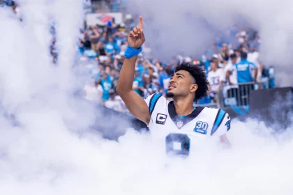 FootballR - NFL - Bryce Young - Ein Footballspieler trägt ein weißes Panthers-Trikot mit der Nummer 9 und blauen und schwarzen Akzenten und ist von Rauch umgeben, während er mit seiner rechten Hand nach oben gestikuliert. Er steht auf einem Spielfeld und die Tribünen im Hintergrund sind voller Fans, von denen einige blau-weiße Trikots tragen. Diese Beschreibung wurde automatisch generiert.
