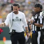 FootballR - NFL - Matt LaFleur, Trainer der Green Bay Packers, trägt ein weißes Langarmshirt mit dem Logo des Teams, ein Headset und eine schwarze Kappe und wirft eine rote Challenge-Flagge. Ein Schiedsrichter in einer schwarz-weiß gestreiften Uniform beobachtet aufmerksam, während sich im Hintergrund verschwommene Footballspieler in Grün und Gold auf dem Feld bewegen. Diese Beschreibung wurde automatisch generiert.