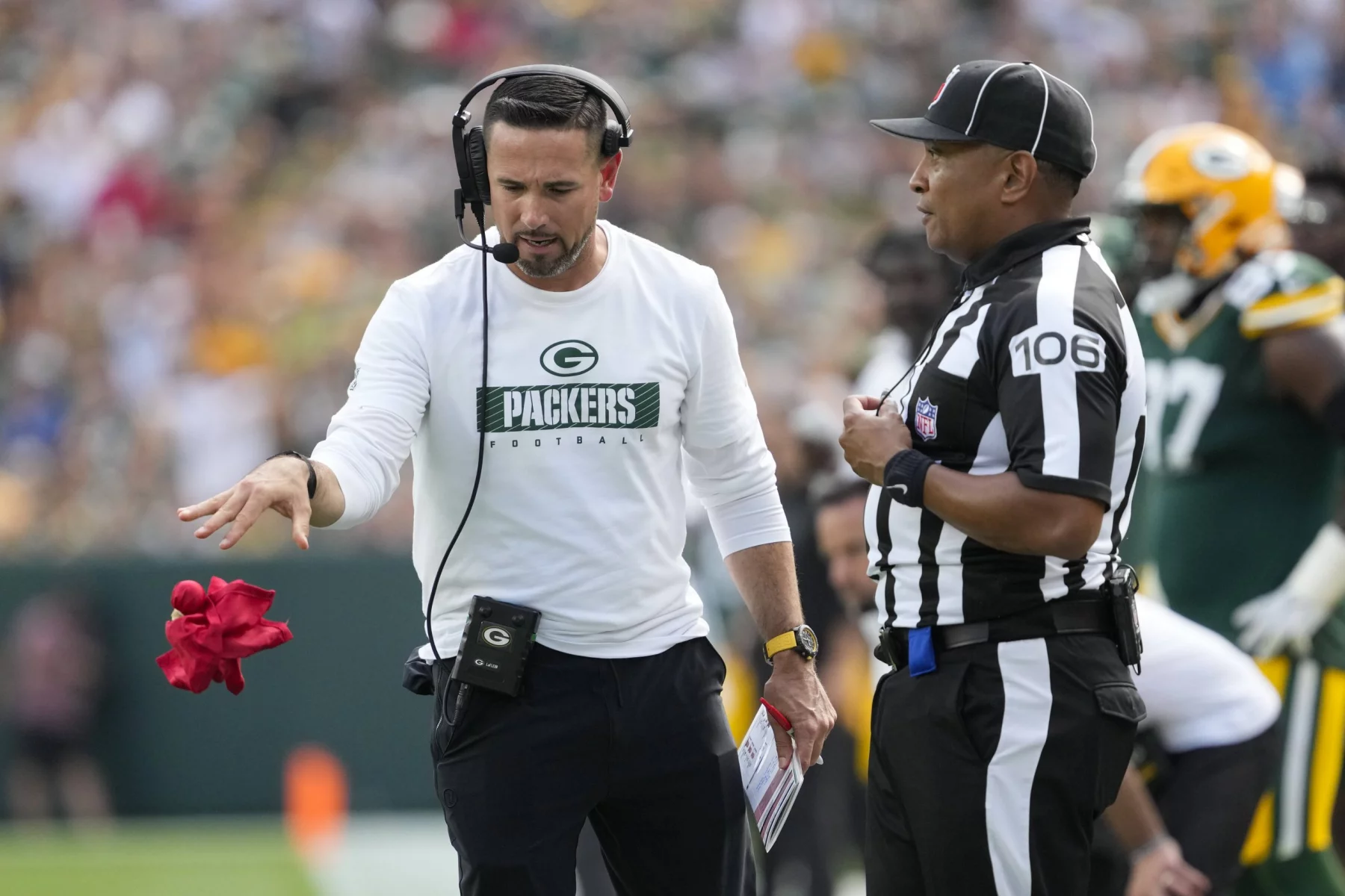 FootballR - NFL - Matt LaFleur, Trainer der Green Bay Packers, trägt ein weißes Langarmshirt mit dem Logo des Teams, ein Headset und eine schwarze Kappe und wirft eine rote Challenge-Flagge. Ein Schiedsrichter in einer schwarz-weiß gestreiften Uniform beobachtet aufmerksam, während sich im Hintergrund verschwommene Footballspieler in Grün und Gold auf dem Feld bewegen. Diese Beschreibung wurde automatisch generiert.