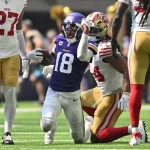 FootballR - NFL - Ein Footballspieler in lila-weißer Uniform, Justin Jefferson, rennt mit dem Ball in der Hand über das Feld. Er wird von gegnerischen Spielern in weiß-roten Uniformen verfolgt. Die Szene spielt sich auf einem Rasenfeld ab, im Hintergrund sind Zuschauer und Cheerleader zu sehen. Diese Beschreibung wurde automatisch generiert.