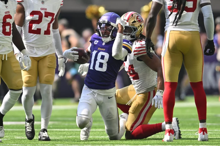FootballR - NFL - Ein Footballspieler in lila-weißer Uniform, Justin Jefferson, rennt mit dem Ball in der Hand über das Feld. Er wird von gegnerischen Spielern in weiß-roten Uniformen verfolgt. Die Szene spielt sich auf einem Rasenfeld ab, im Hintergrund sind Zuschauer und Cheerleader zu sehen. Diese Beschreibung wurde automatisch generiert.
