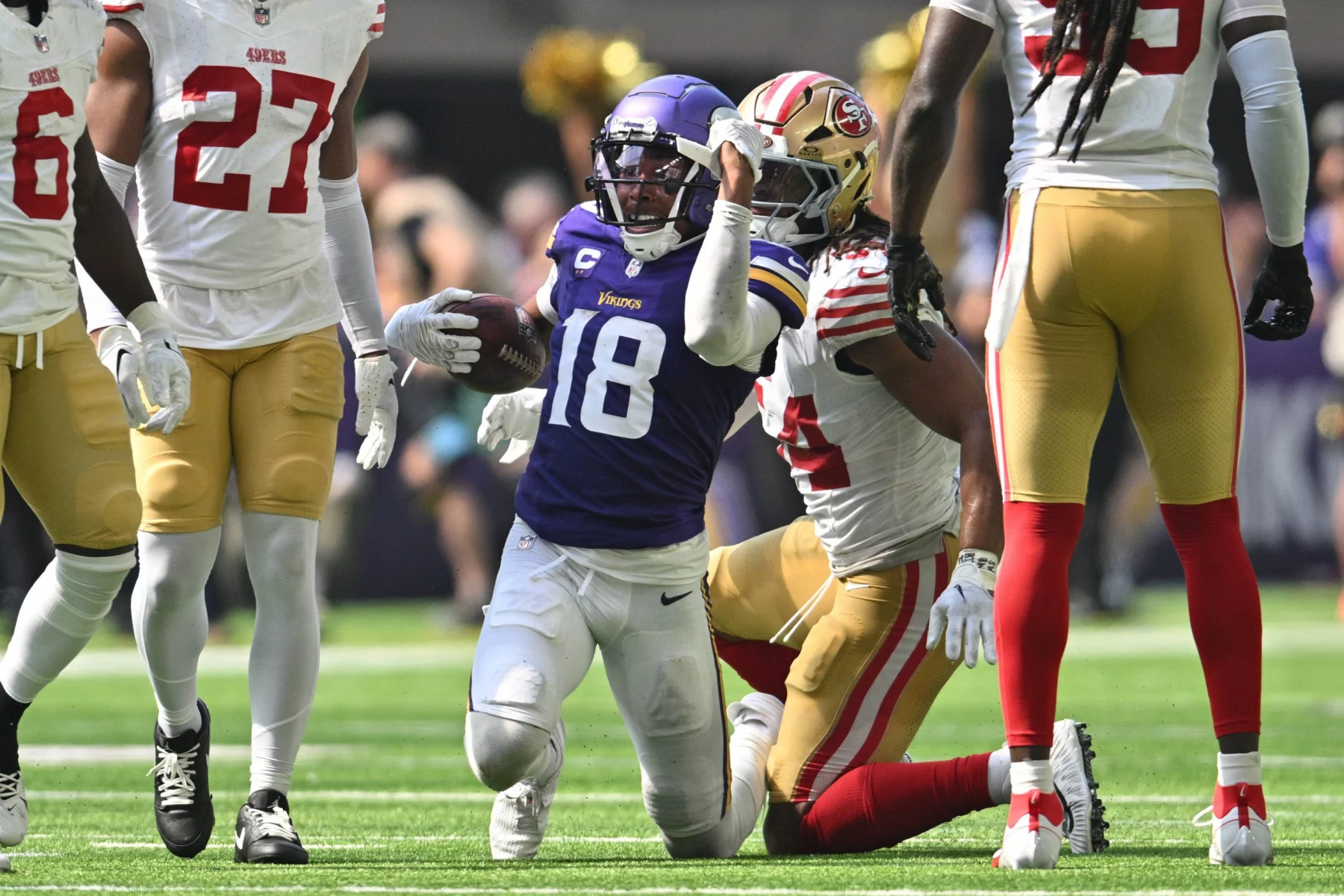 FootballR - NFL - Ein Footballspieler in lila-weißer Uniform, Justin Jefferson, rennt mit dem Ball in der Hand über das Feld. Er wird von gegnerischen Spielern in weiß-roten Uniformen verfolgt. Die Szene spielt sich auf einem Rasenfeld ab, im Hintergrund sind Zuschauer und Cheerleader zu sehen. Diese Beschreibung wurde automatisch generiert.