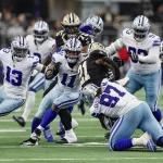 FootballR - NFL - Footballspieler der Dallas Cowboys und der New Orleans Saints spielen. Die Nummern 11 - Micah Parsons - und 13 der Cowboys machen einen Angriff, während die Nummer 97 angegriffen wird. Saints-Spieler in schwarz-goldenen Uniformen versuchen, sie aufzuhalten. Die Action spielt sich auf einem grünen Feld ab, im Hintergrund sind Zuschauer und die Seitenlinie unscharf. Diese Beschreibung wurde automatisch generiert.
