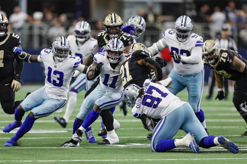 FootballR - NFL - Footballspieler der Dallas Cowboys und der New Orleans Saints spielen. Die Nummern 11 - Micah Parsons - und 13 der Cowboys machen einen Angriff, während die Nummer 97 angegriffen wird. Saints-Spieler in schwarz-goldenen Uniformen versuchen, sie aufzuhalten. Die Action spielt sich auf einem grünen Feld ab, im Hintergrund sind Zuschauer und die Seitenlinie unscharf. Diese Beschreibung wurde automatisch generiert.