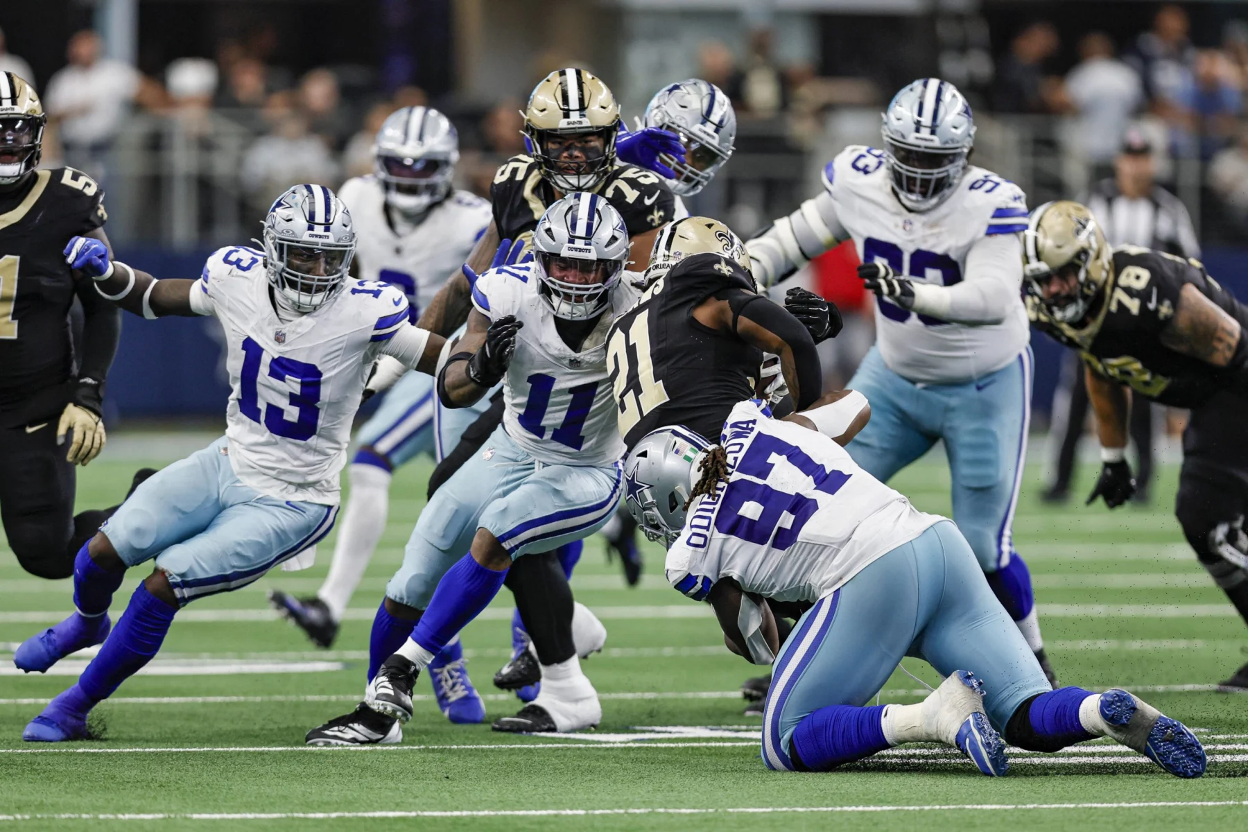 FootballR - NFL - Footballspieler der Dallas Cowboys und der New Orleans Saints spielen. Die Nummern 11 - Micah Parsons - und 13 der Cowboys machen einen Angriff, während die Nummer 97 angegriffen wird. Saints-Spieler in schwarz-goldenen Uniformen versuchen, sie aufzuhalten. Die Action spielt sich auf einem grünen Feld ab, im Hintergrund sind Zuschauer und die Seitenlinie unscharf. Diese Beschreibung wurde automatisch generiert.