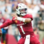FootballR - NFL Woche 2 - Ein Spieler der Arizona Cardinals in einem roten Trikot mit der Nummer 1, Kyler Murray, bereitet sich während eines Spiels darauf vor, einen Football zu werfen. Der Spieler ist in Bewegung, hat seinen rechten Arm nach hinten gestreckt und den Blick nach unten gerichtet. Er trägt einen weißen Helm mit dem Logo des Teams. Im Hintergrund sind verschwommene Zuschauer zu sehen. Diese Beschreibung wurde automatisch generiert.