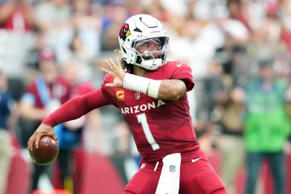 FootballR - NFL Woche 2 - Ein Spieler der Arizona Cardinals in einem roten Trikot mit der Nummer 1, Kyler Murray, bereitet sich während eines Spiels darauf vor, einen Football zu werfen. Der Spieler ist in Bewegung, hat seinen rechten Arm nach hinten gestreckt und den Blick nach unten gerichtet. Er trägt einen weißen Helm mit dem Logo des Teams. Im Hintergrund sind verschwommene Zuschauer zu sehen. Diese Beschreibung wurde automatisch generiert.