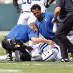 FootballR - NFL - Ein verletzter Colts-Defense-Spieler in weiß-blauer Uniform liegt auf dem Feld, während sich drei Trainer in schwarz-blauen Hemden um ihn kümmern. Während er sich offensichtlich schmerzerfüllt den Kopf hält, untersuchen und trösten sie ihn. Die Szene spielt sich während eines Tagesspiels ab, und man macht sich Sorgen um DeForest Buckner, der möglicherweise auf die Verletztenliste kommt. Diese Beschreibung wurde automatisch generiert.