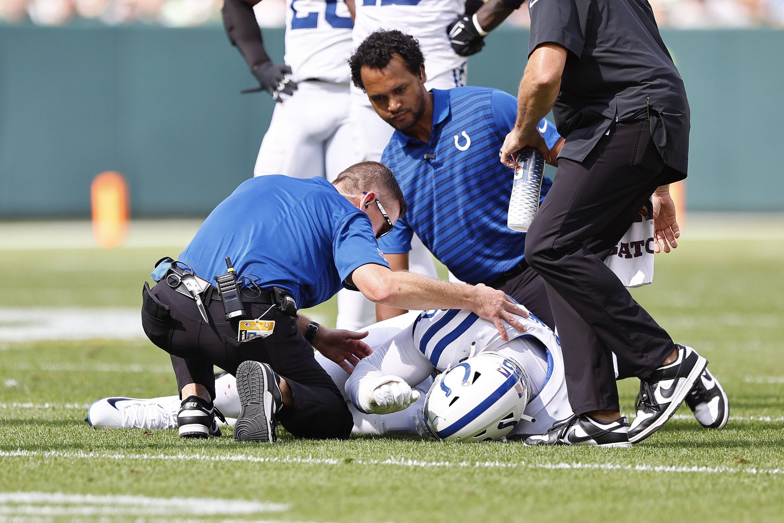 FootballR - NFL - Ein verletzter Colts-Defense-Spieler in weiß-blauer Uniform liegt auf dem Feld, während sich drei Trainer in schwarz-blauen Hemden um ihn kümmern. Während er sich offensichtlich schmerzerfüllt den Kopf hält, untersuchen und trösten sie ihn. Die Szene spielt sich während eines Tagesspiels ab, und man macht sich Sorgen um DeForest Buckner, der möglicherweise auf die Verletztenliste kommt. Diese Beschreibung wurde automatisch generiert.